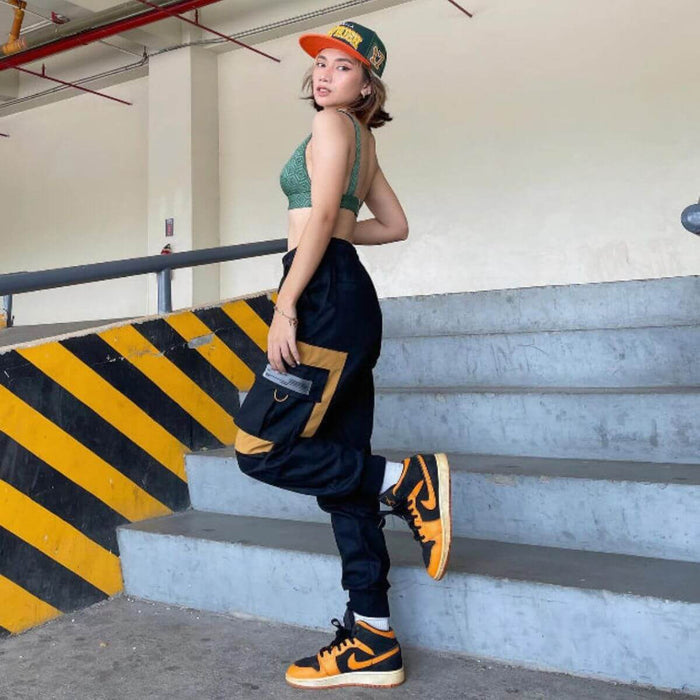 Woman wearing a green crop top, black cargo pants, and orange sneakers, posing on stairs in an industrial setting.