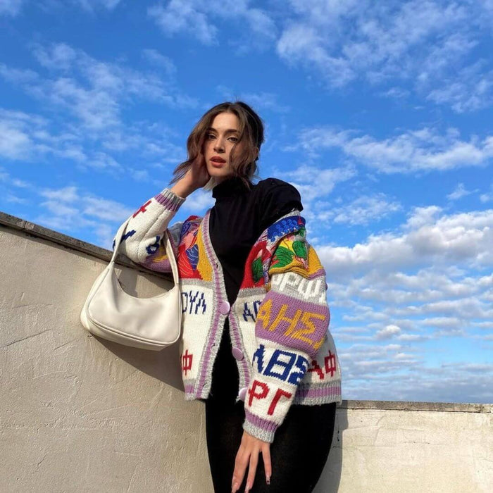 Woman wearing a colorful knit cardigan and holding a white handbag, posing against a bright blue sky.