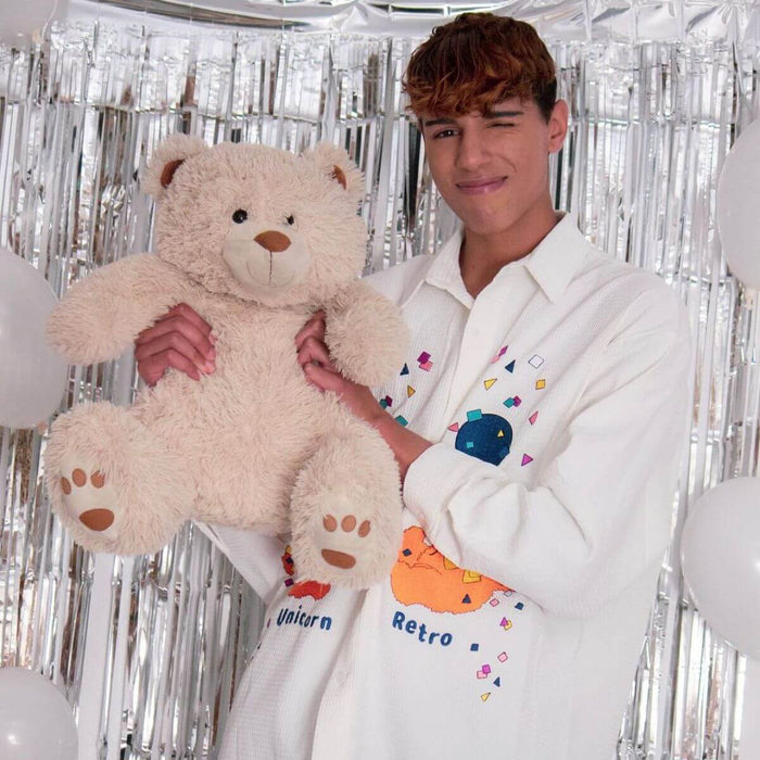Man wearing a white shirt with colorful prints, holding a plush teddy bear, standing in front of a silver fringe backdrop.