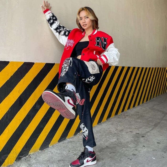 Woman wearing a red and black varsity jacket, graphic pants, and sneakers, leaning against a wall with yellow and black stripes.